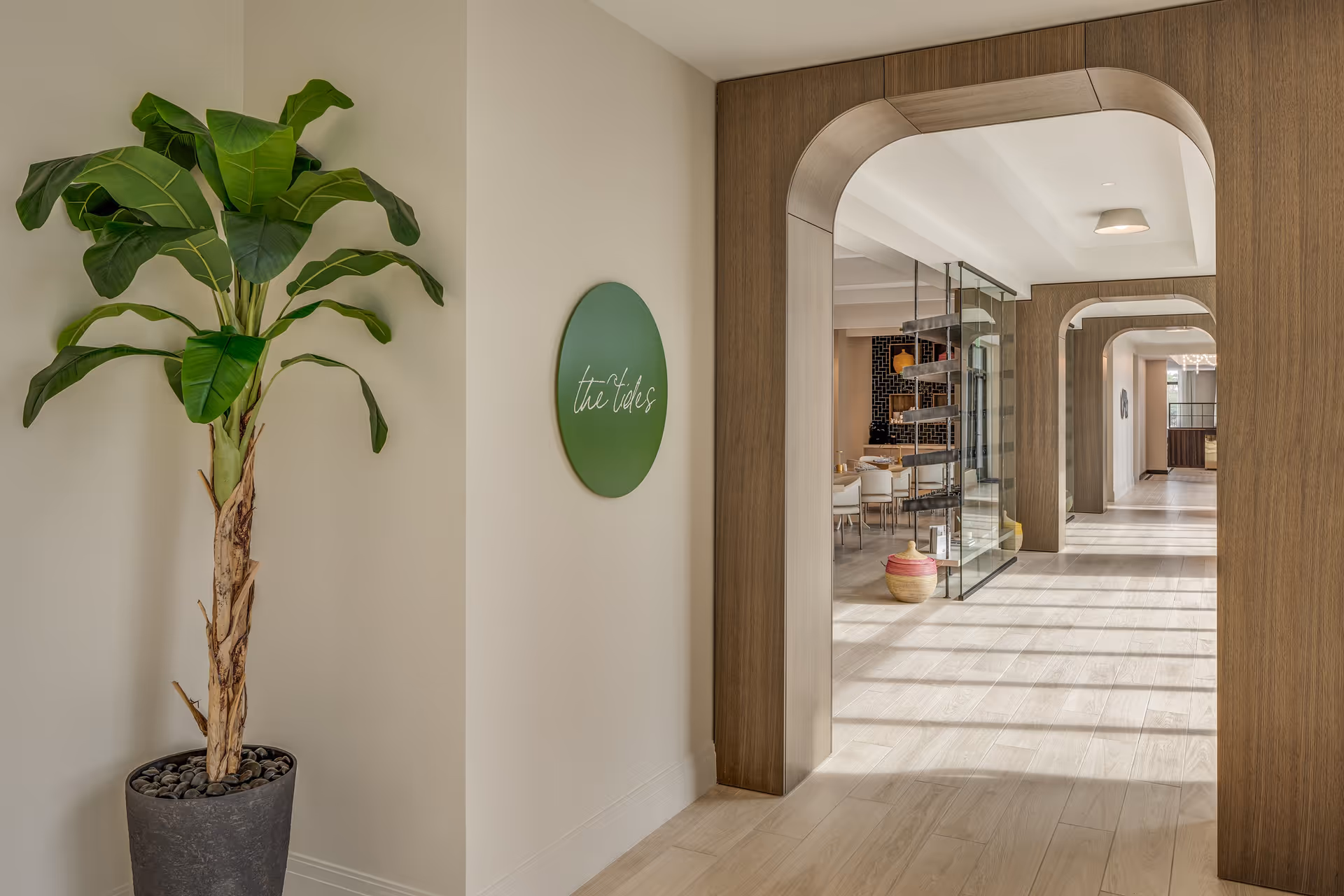 A bright hallway with light wood flooring and arched wooden door frames. On the left side, there is a tall potted plant with large green leaves and a round green sign on the wall that reads 'the tides'. The hallway leads to a dining area with tables and chairs visible through glass partitions.