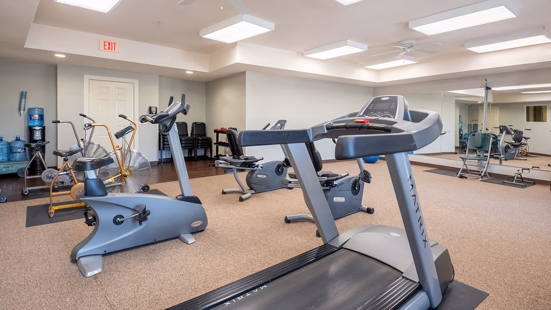A fitness room in a senior living facility featuring exercise equipment including a treadmill, stationary bikes, and an air bike. The room has beige carpet, white walls, a ceiling fan, and a large mirror on one wall. There is a water dispenser and stacked chairs in the background.