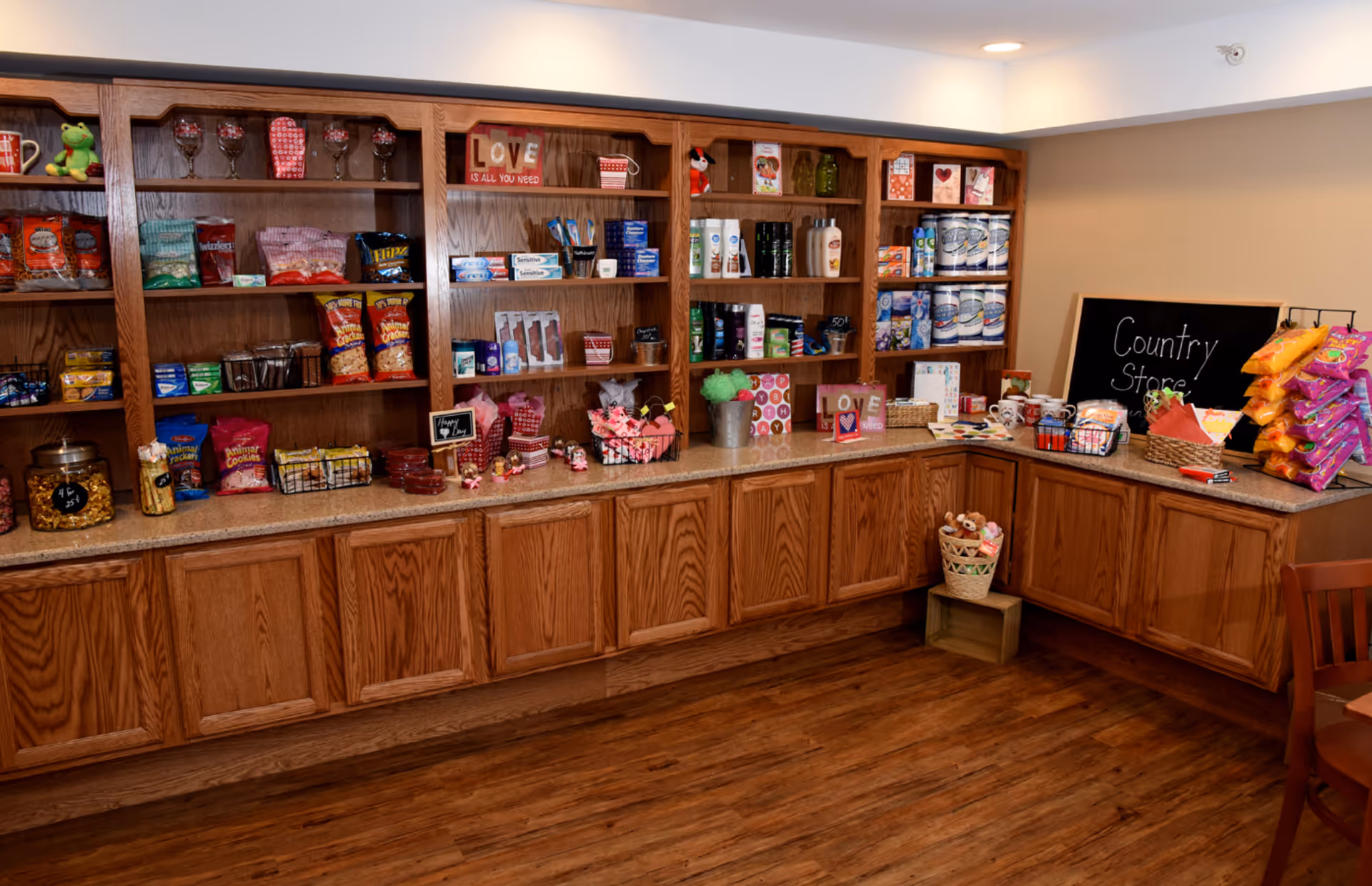 Interior view of a country store area in a senior living facility with wooden shelves and cabinets stocked with snacks, personal care items, and small gift items. A chalkboard sign reads 'Country Store'. The floor is wooden and there is a wooden chair partially visible on the right.