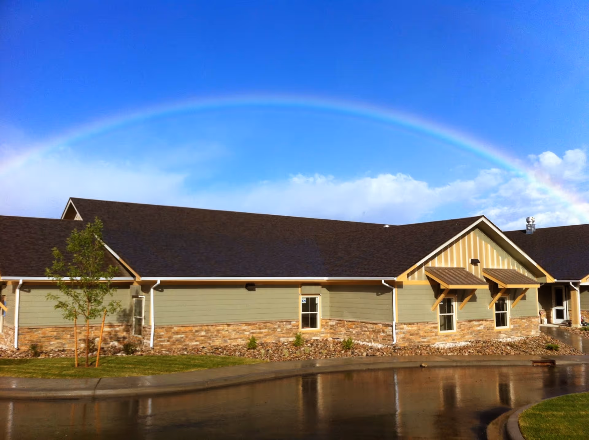 Exterior of a single-story building with a rainbow arching across a clear blue sky.