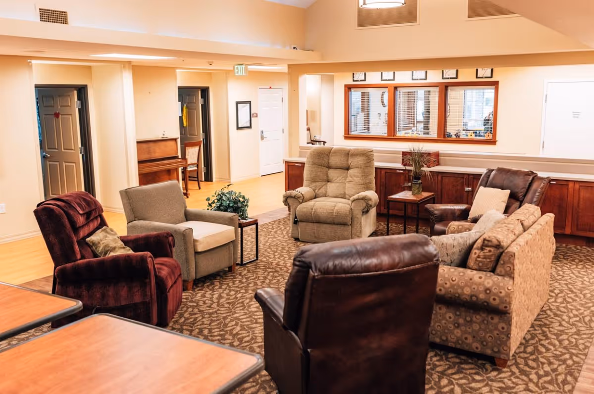 A cozy seating area in an assisted living facility with various armchairs and a sofa arranged in a circle on a patterned carpet. The room has light-colored walls, wooden cabinets along one wall, a piano in the background, and a large window with blinds. There are also tables with plants and decorative items.