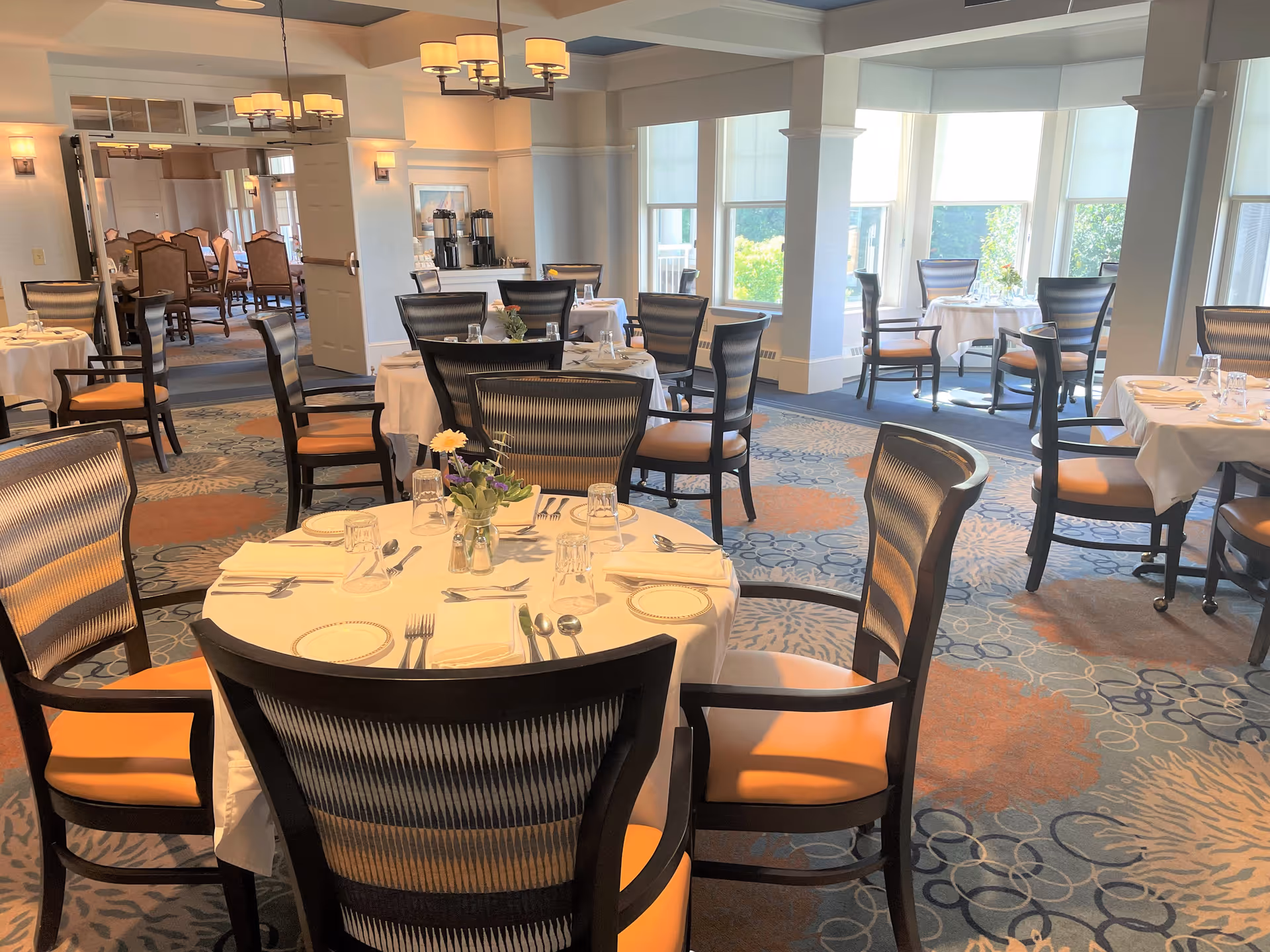 A bright dining room with round tables covered in white tablecloths, each set with plates, glasses, and silverware. The chairs have dark frames with orange cushions. Large windows allow natural light to fill the room, and there are floral centerpieces on the tables. In the background, there is another dining area visible through an open doorway.