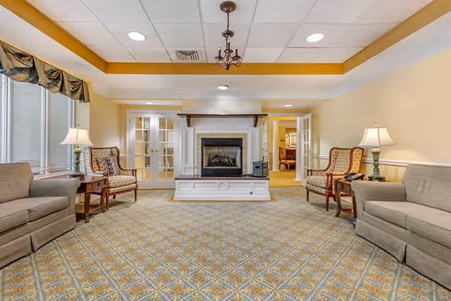 A spacious living room with patterned carpet, two beige sofas, two upholstered armchairs, wooden side tables with lamps, and a white fireplace in the center. The room has large windows with valances on the left and recessed lighting in the ceiling.