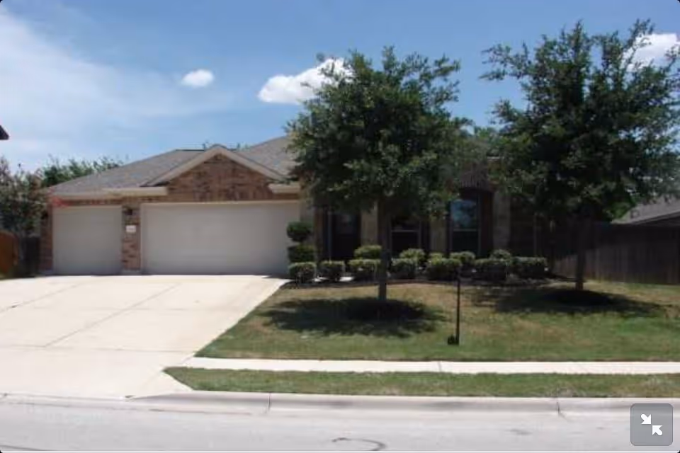 Front exterior view of a single-story brick house with a three-car garage, a concrete driveway, two trees, and a lawn with trimmed bushes.