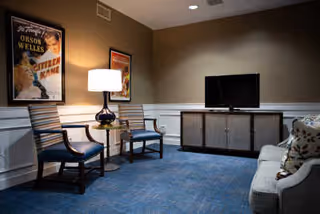 A cozy living room area with two wooden chairs with striped cushions, a small round side table with a lamp, a flat-screen TV on a cabinet, and a sofa with patterned pillows. The walls are decorated with framed vintage movie posters.