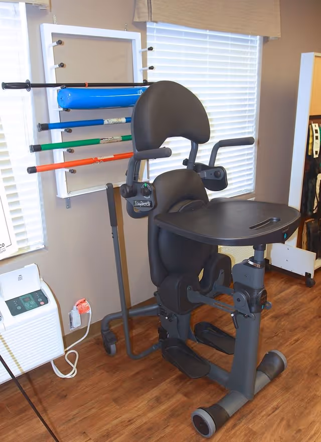 A black exercise machine with a padded seat, backrest, armrests, and a tray in front, positioned on a wooden floor in a room with windows covered by blinds. Behind the machine, there is a rack mounted on the wall holding various colored exercise rods. A white electronic device with a power cord is placed on the floor to the left.
