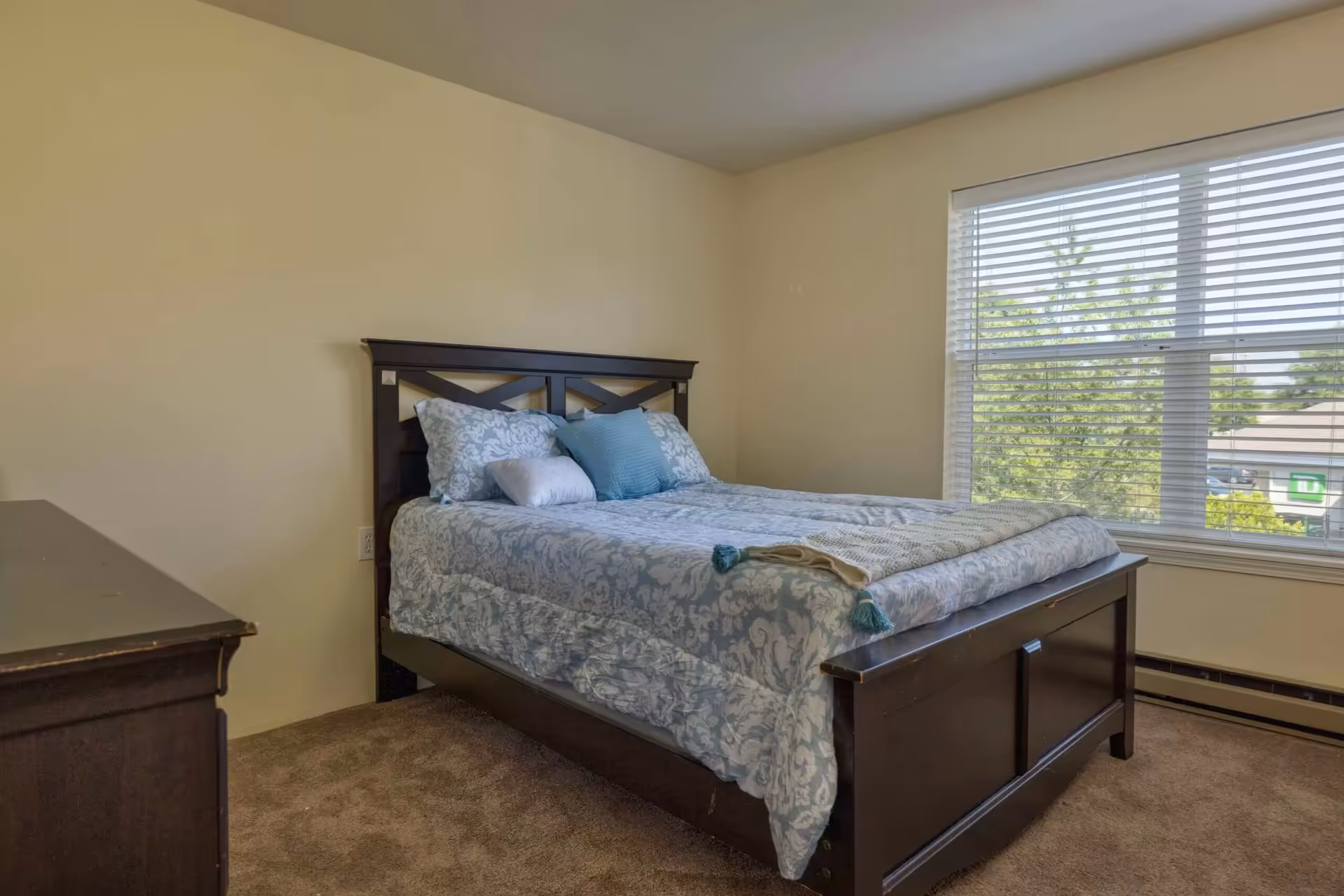 A bedroom with a dark wooden bed frame and a neatly made bed with light blue and white patterned bedding and pillows. There is a large window with white blinds letting in natural light, and a dark wooden dresser is partially visible on the left side of the image. The walls are light-colored and the floor is carpeted.