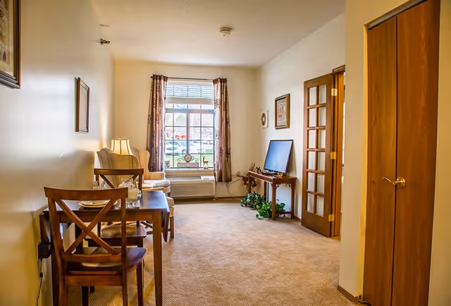 A cozy living room area with beige carpet and cream-colored walls. There is a wooden dining table with two chairs on the left side, set with plates and glasses. A comfortable armchair with a floor lamp is positioned near a large window with patterned curtains. On the right side, a flat-screen TV sits on a wooden stand with a green plant underneath. A wooden door with glass panels and a closed wooden closet door are visible on the right wall.