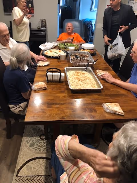 A group of elderly people sitting and standing around a wooden dining table with various dishes of food, including a large tray of rice, salad, and other prepared meals. The setting appears to be a communal dining area inside a facility.