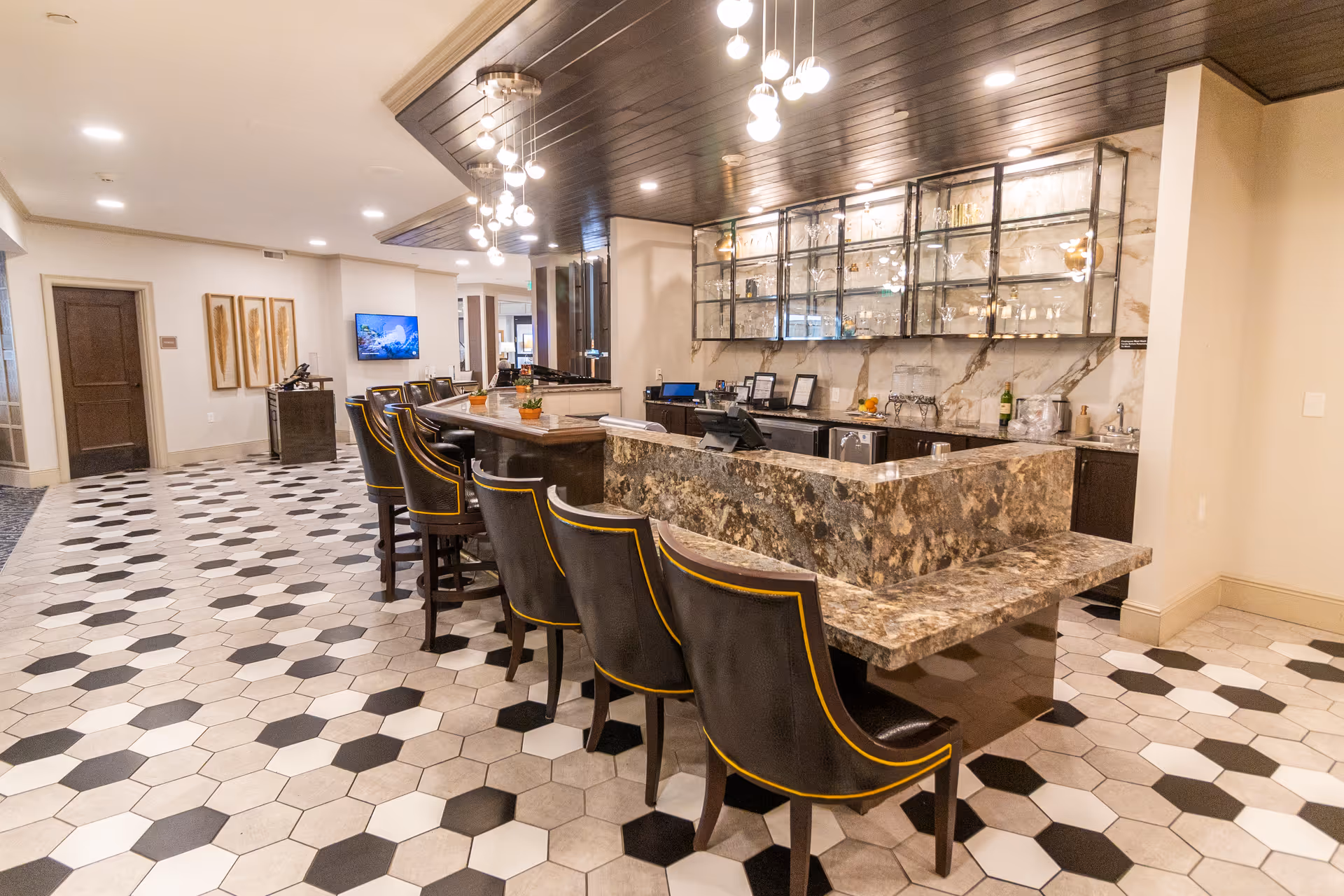 Interior view of a modern bar area in a senior living facility with a marble countertop, black leather bar stools with yellow trim, hexagonal floor tiles in white, gray, and black, glass shelves with glassware, and warm lighting fixtures hanging from a wooden ceiling.