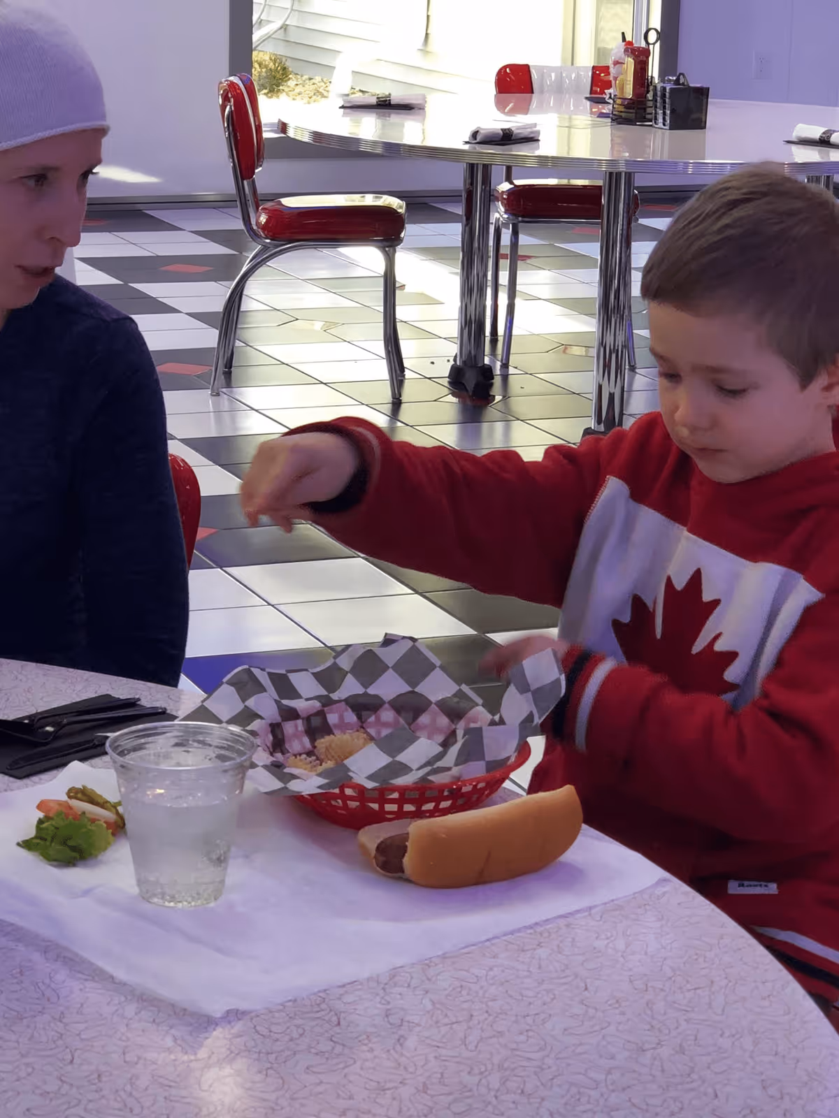 A young boy in a red and white sweater with a maple leaf design is sitting at a table with a woman wearing a light purple beanie. The boy is reaching into a red basket lined with checkered paper that contains waffle fries. On the table, there is a hot dog, a plastic cup of water, and some lettuce and tomato. The background shows a retro-style diner with red and chrome chairs and a black, white, and red tiled floor.