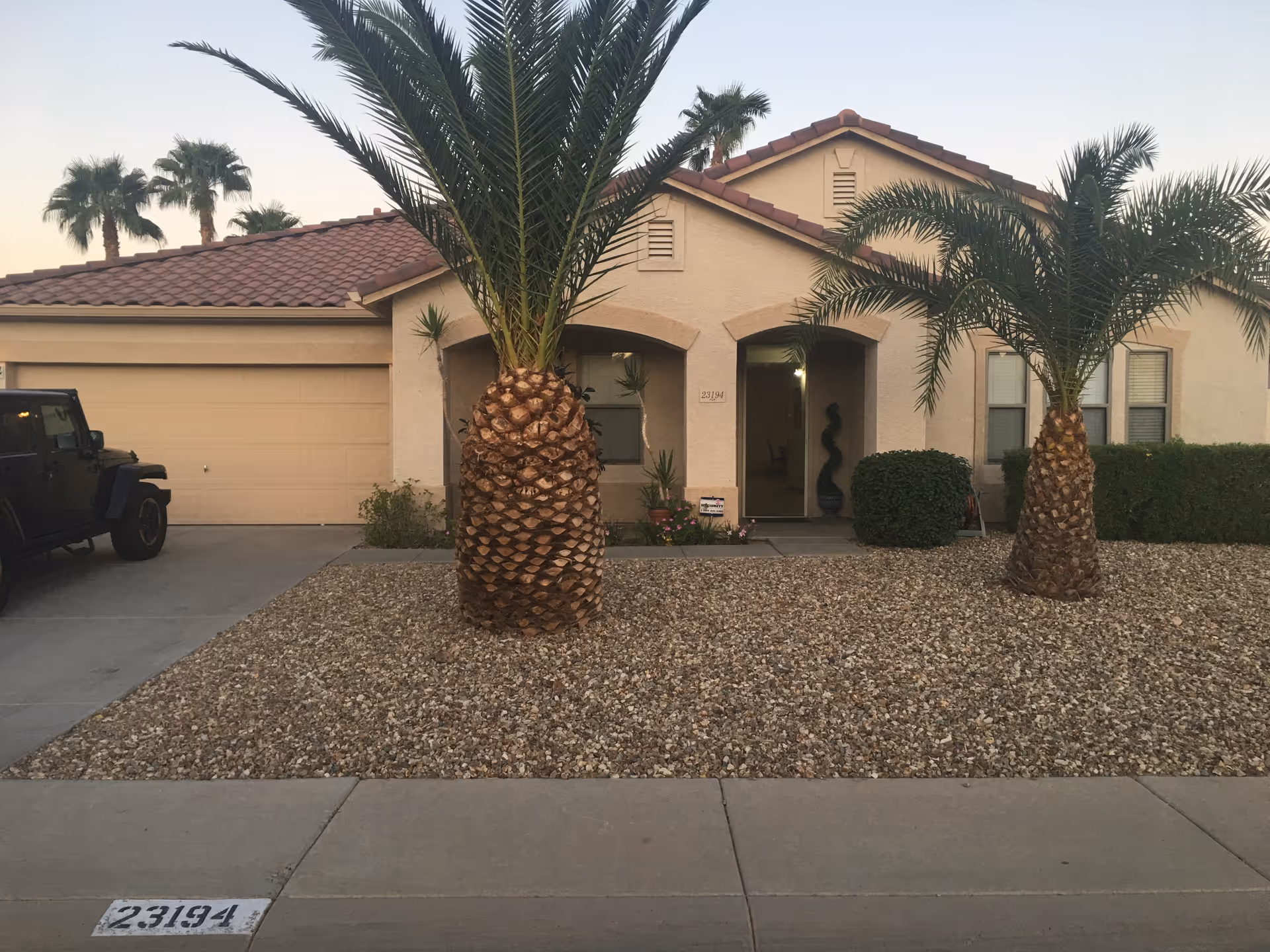 Front exterior of a single-story stucco house with two palm trees in a gravel front yard and an attached garage.