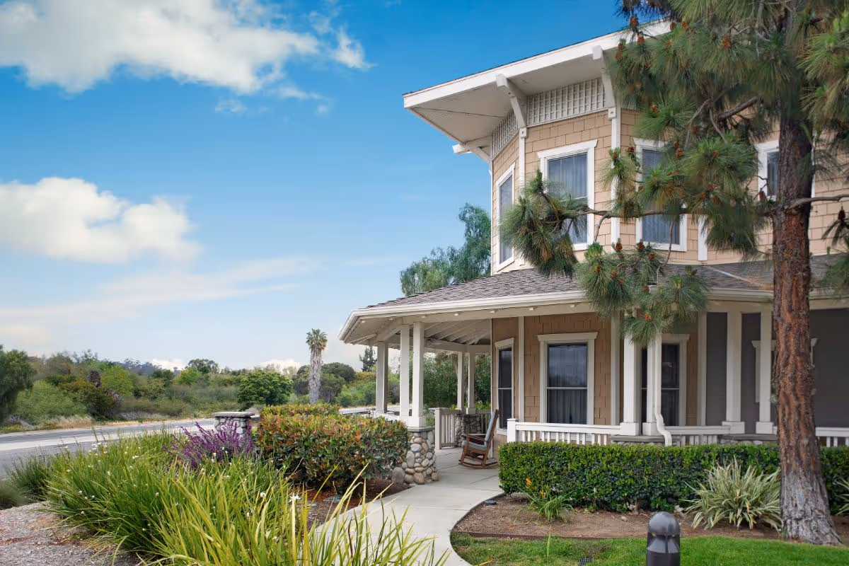 Exterior view of a two-story residential building with beige siding and white trim, featuring a covered porch with a rocking chair. The surrounding area includes landscaped bushes, trees, and a clear blue sky with some clouds.