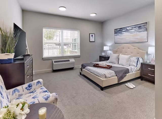Well-lit bedroom with a tufted bed, nightstands, a dresser with TV, and a window with blinds.