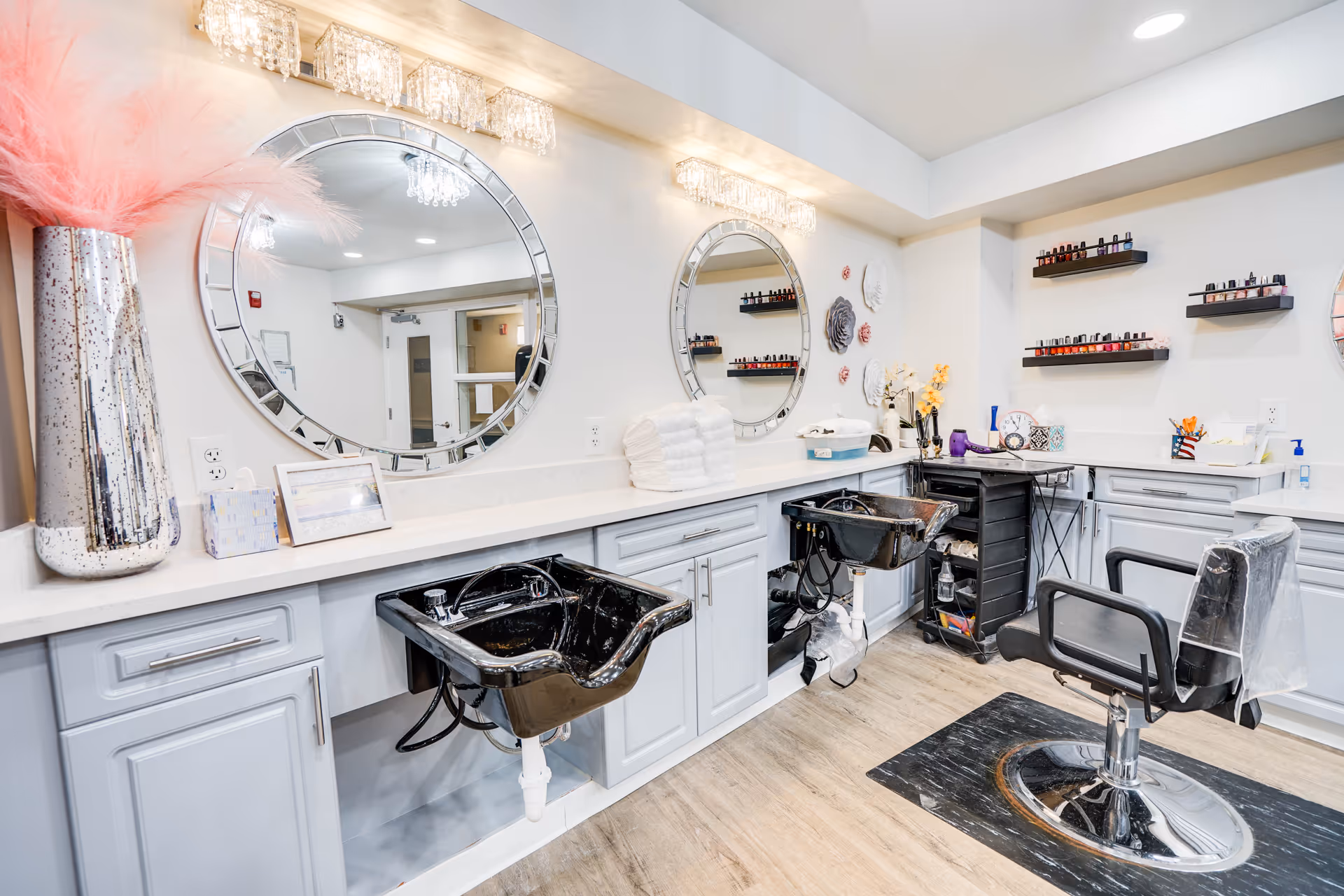 A bright and clean salon area with two black hair washing sinks, a black salon chair on a protective mat, two large round mirrors with decorative frames, shelves with nail polish bottles, and a countertop with towels and various salon supplies. The room has light-colored walls and wood flooring.
