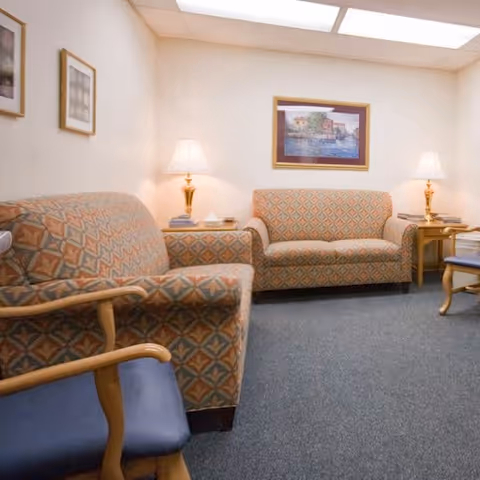 A cozy seating area in a senior living facility with two patterned sofas, a wooden armchair with blue cushion, two side tables each with a lamp, and framed artwork on the walls under bright ceiling lights.