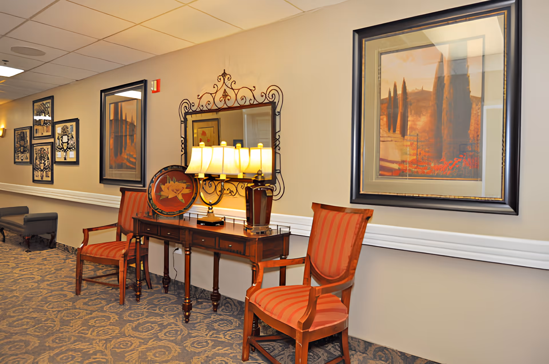 A decorated interior hallway seating area with two striped wooden chairs, a console table holding lamps and a mirror, and framed artwork on the wall.