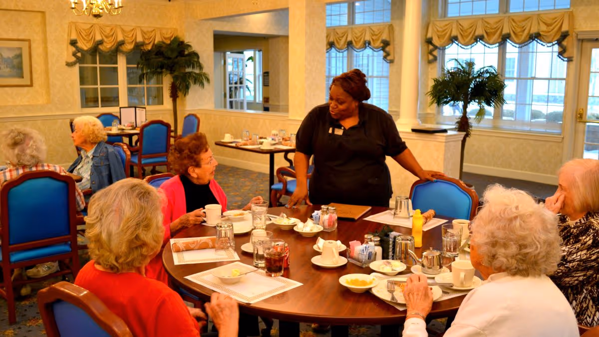 A group of elderly women seated around a dining table in a well-lit room with large windows and blue chairs. A staff member in a black uniform is standing and engaging with the women. The table is set with cups, plates, and condiments, indicating a meal setting.