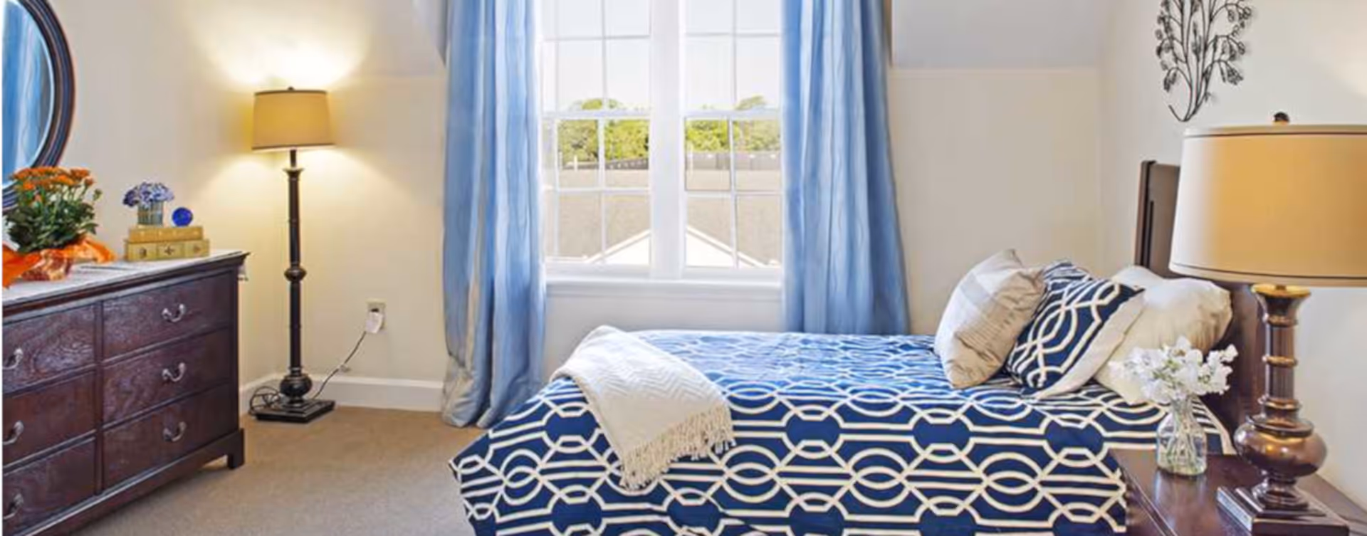 Sunlit bedroom with a bed dressed in navy geometric bedding, a wooden dresser, floor lamp, bedside table with lamp and flowers, and a window with blue curtains.