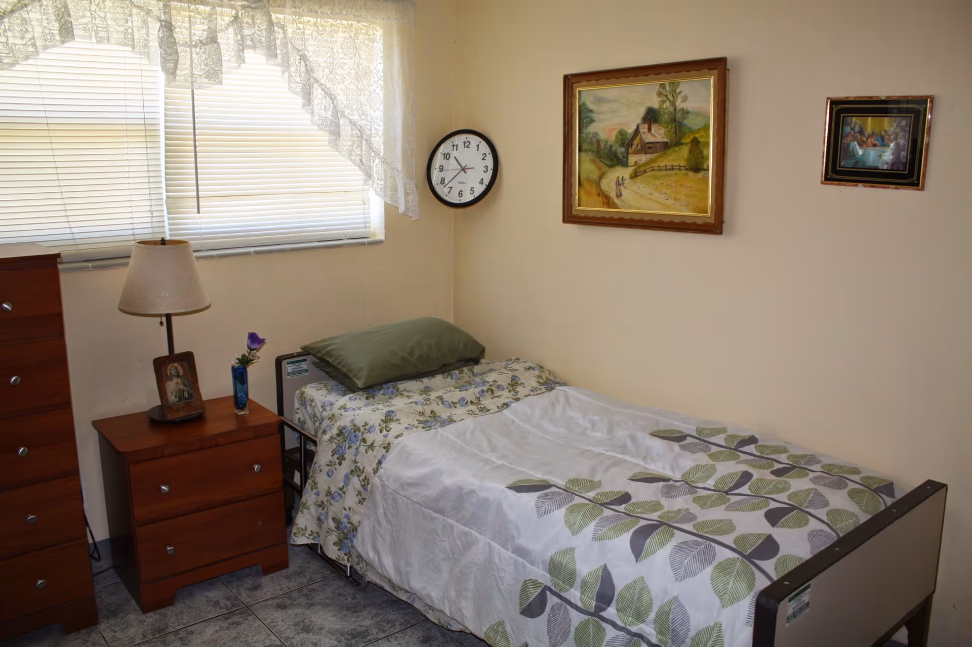 A small bedroom with a single bed covered with a white and green leaf-patterned blanket and a floral sheet. There is a green pillow on the bed. Next to the bed is a wooden nightstand with a lamp, a framed photo, and a small vase with a purple flower. A wooden dresser is beside the nightstand. The room has a window with blinds and lace curtains. On the wall above the bed are two framed paintings and a round clock showing the time as 8:10.