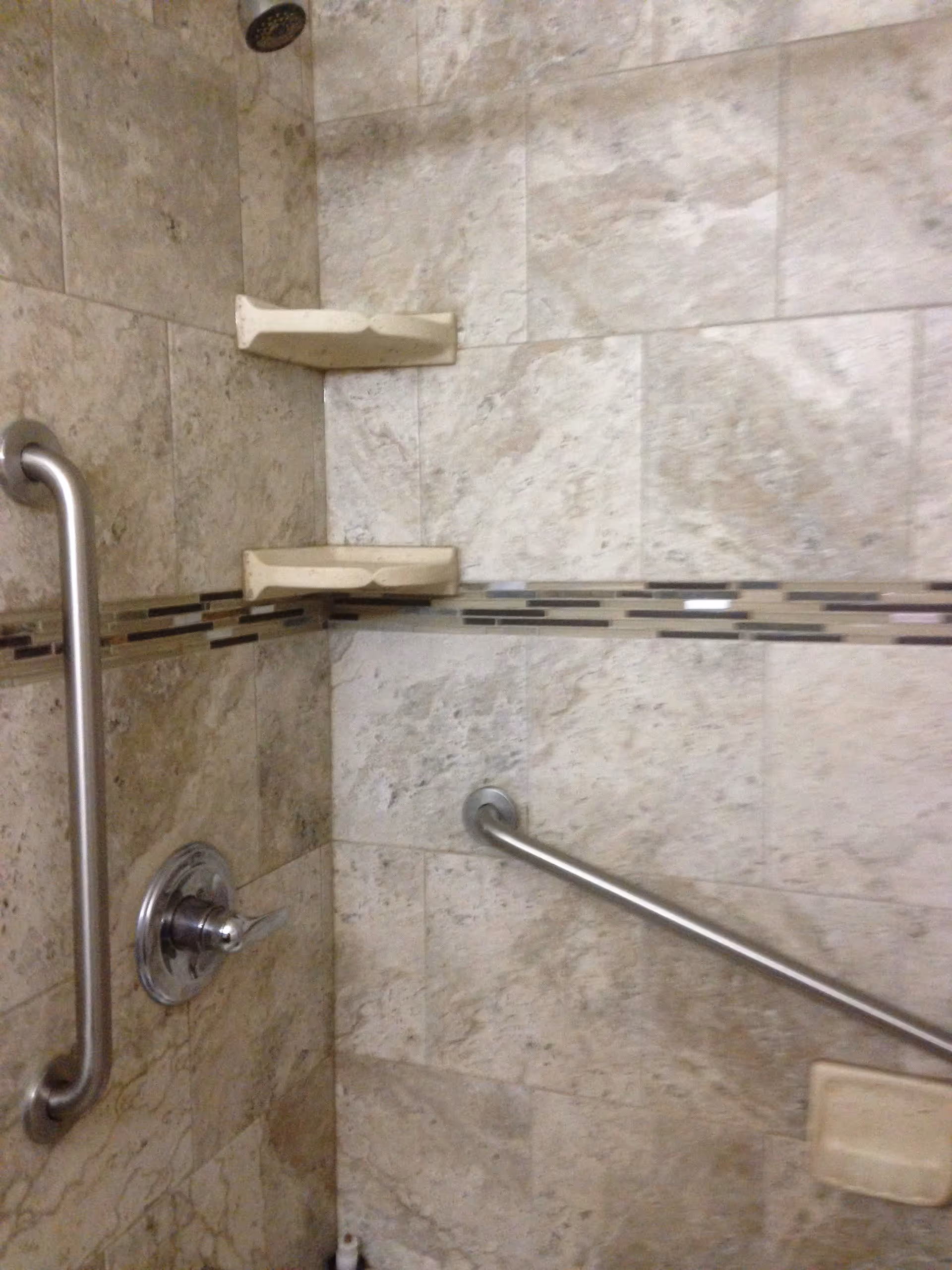 A tiled shower area with beige and brown stone tiles, two corner soap holders, a metal shower handle, and two stainless steel grab bars for support.
