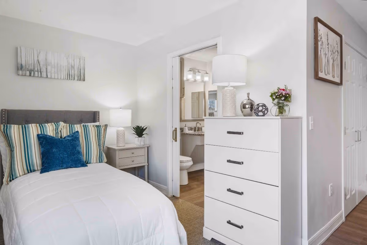 A cozy bedroom with a neatly made bed featuring striped pillows and a blue accent pillow. Next to the bed is a small nightstand with a lamp and a small plant. A white dresser with four drawers stands against the wall, topped with a lamp, decorative items, and a small vase of flowers. An open door reveals a bathroom with a toilet, sink, and mirror. The walls are painted white, and there is a framed artwork on the wall near the dresser.
