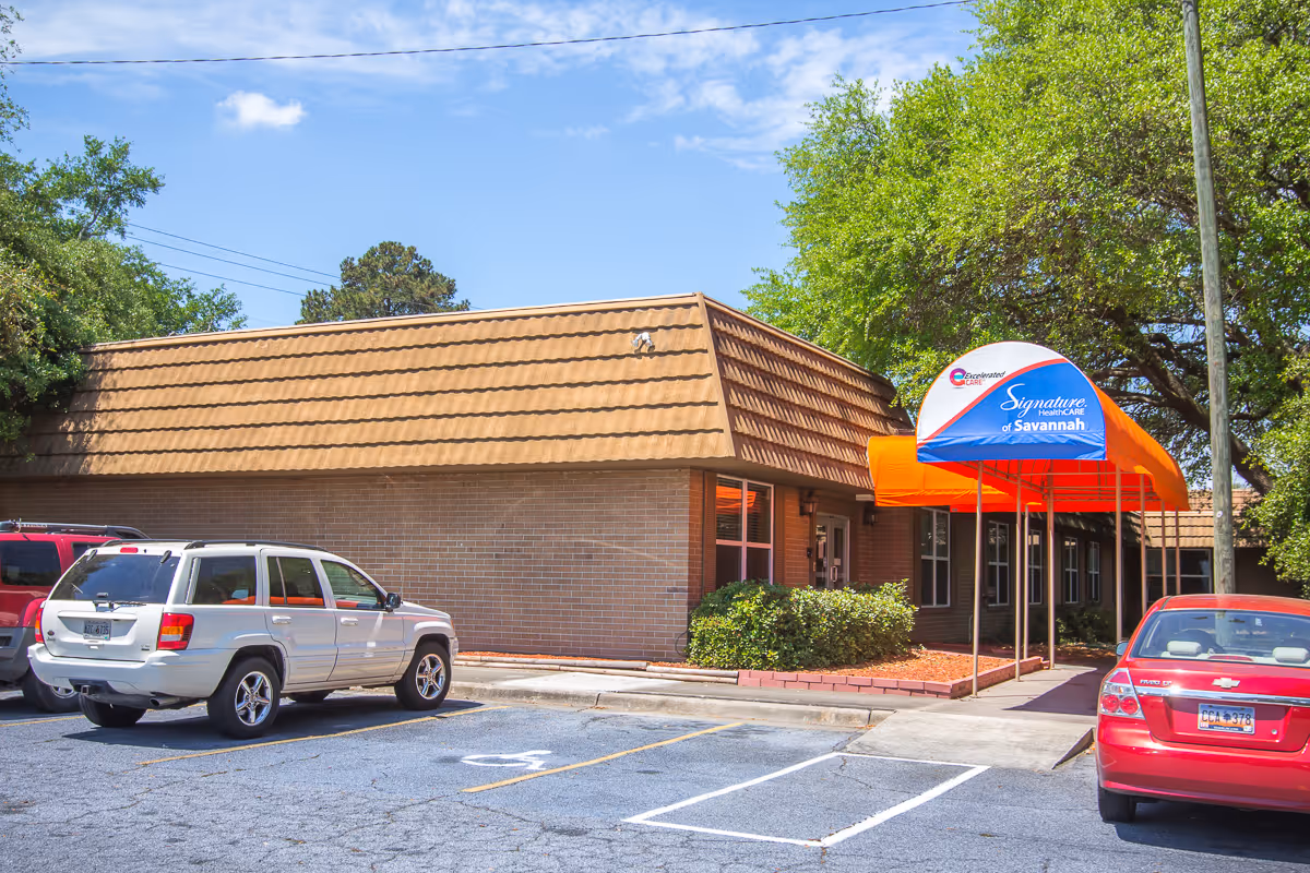 Exterior front of a single-story brick senior care building with an orange entry canopy and a small parking lot with parked cars.