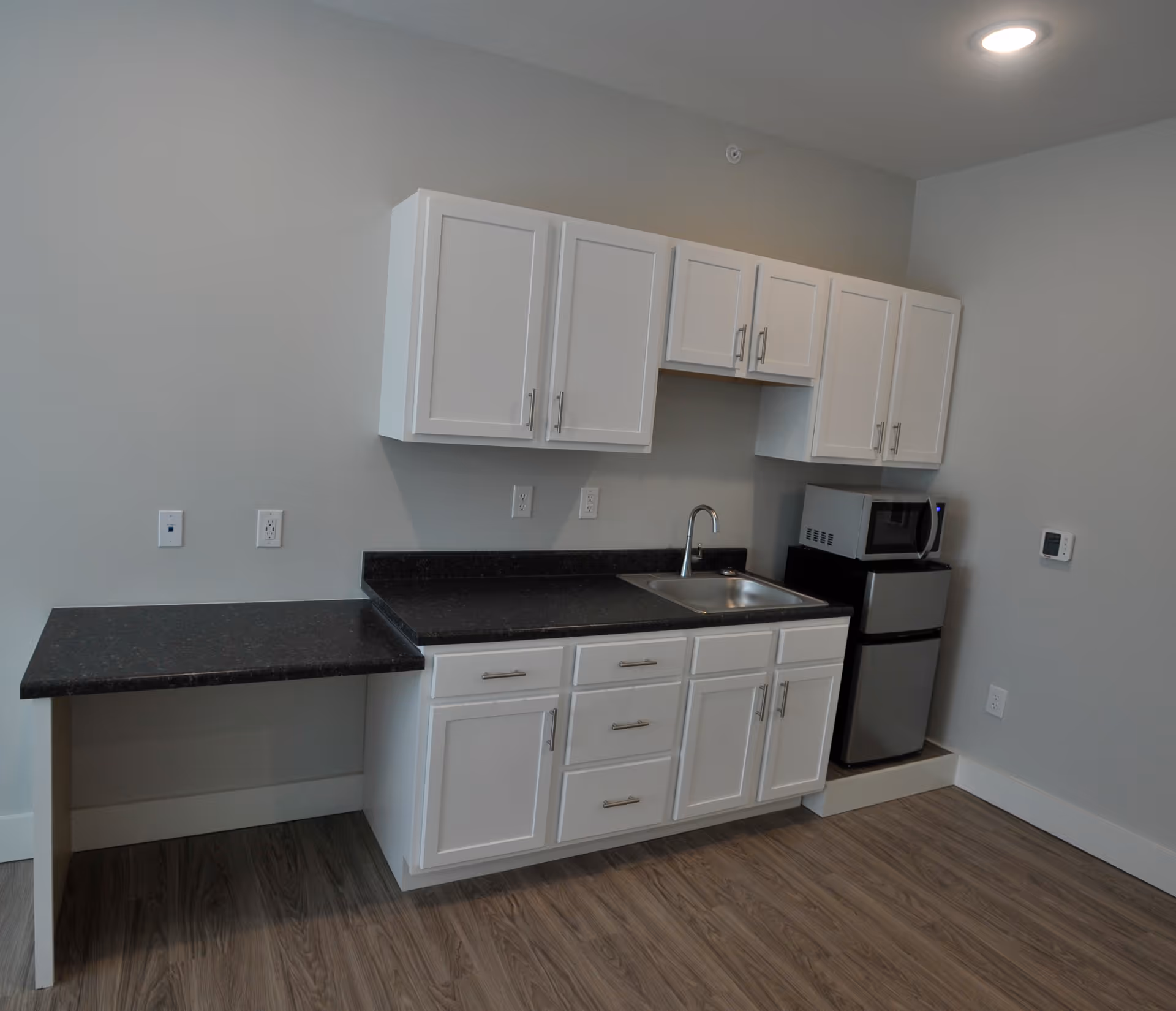 A small kitchenette area with white cabinets, a black countertop, a stainless steel sink, a microwave, and a mini refrigerator. The floor has wood-like flooring and the walls are painted light gray.