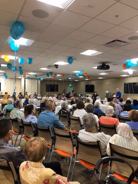 A large group of seniors seated in rows in a decorated community room listening to speakers at the front, with teal and orange balloons hanging from the ceiling.