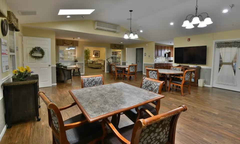 Well-lit communal dining room with several tables and chairs, a wall-mounted TV, and an adjoining kitchenette and seating area.