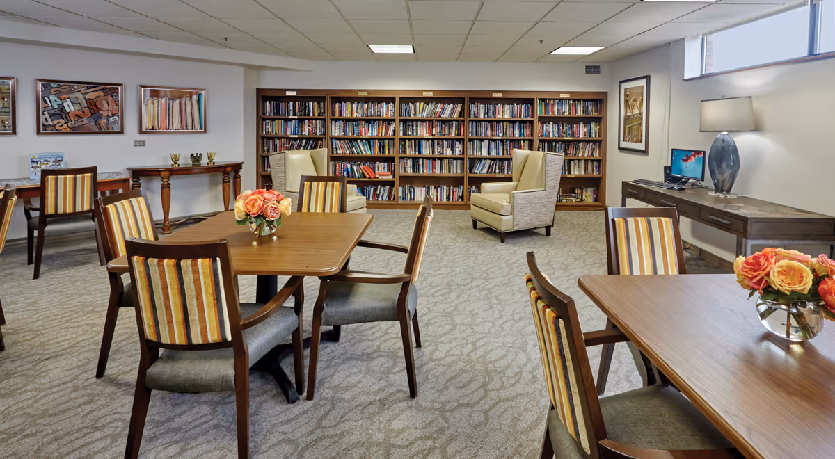 A cozy senior living common area with multiple wooden tables and striped cushioned chairs. There are flower arrangements on the tables, a large bookshelf filled with books along the back wall, and two beige armchairs in front of the bookshelf. A desk with a computer and a lamp is positioned near a window on the right side. The room is carpeted and decorated with framed artwork on the walls.