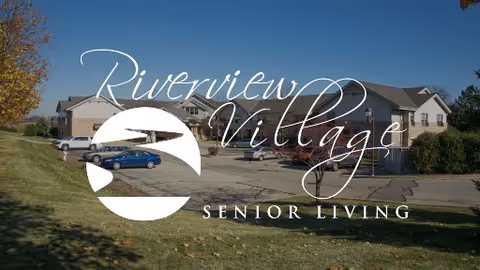 Exterior view of a two-story senior living building with parked cars and a large 'Riverview Village Senior Living' logo overlay.