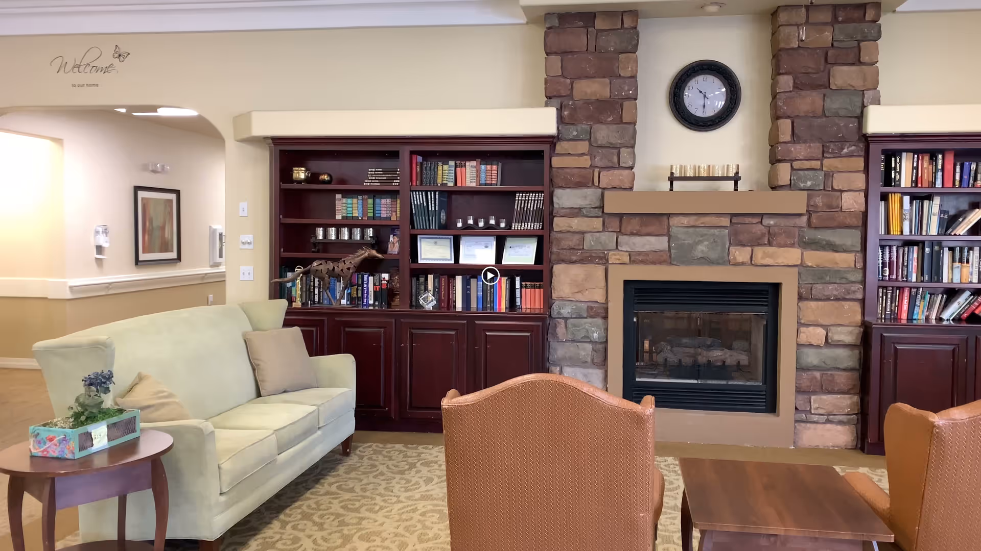 A cozy living room area in an assisted living facility featuring a light green sofa with beige pillows, a wooden side table with a tissue box and small plant, two brown armchairs, a wooden coffee table, built-in dark wood bookshelves filled with books and decorative items, and a stone fireplace with a clock above it.