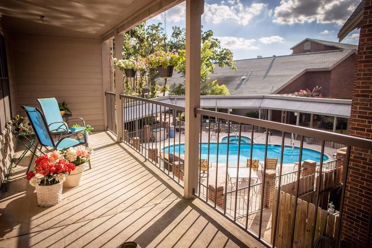 A sunny balcony with two blue chairs, several potted flowers, and hanging flower baskets overlooking a swimming pool and patio area with tables and chairs. The scene includes a view of nearby buildings and trees under a partly cloudy sky.