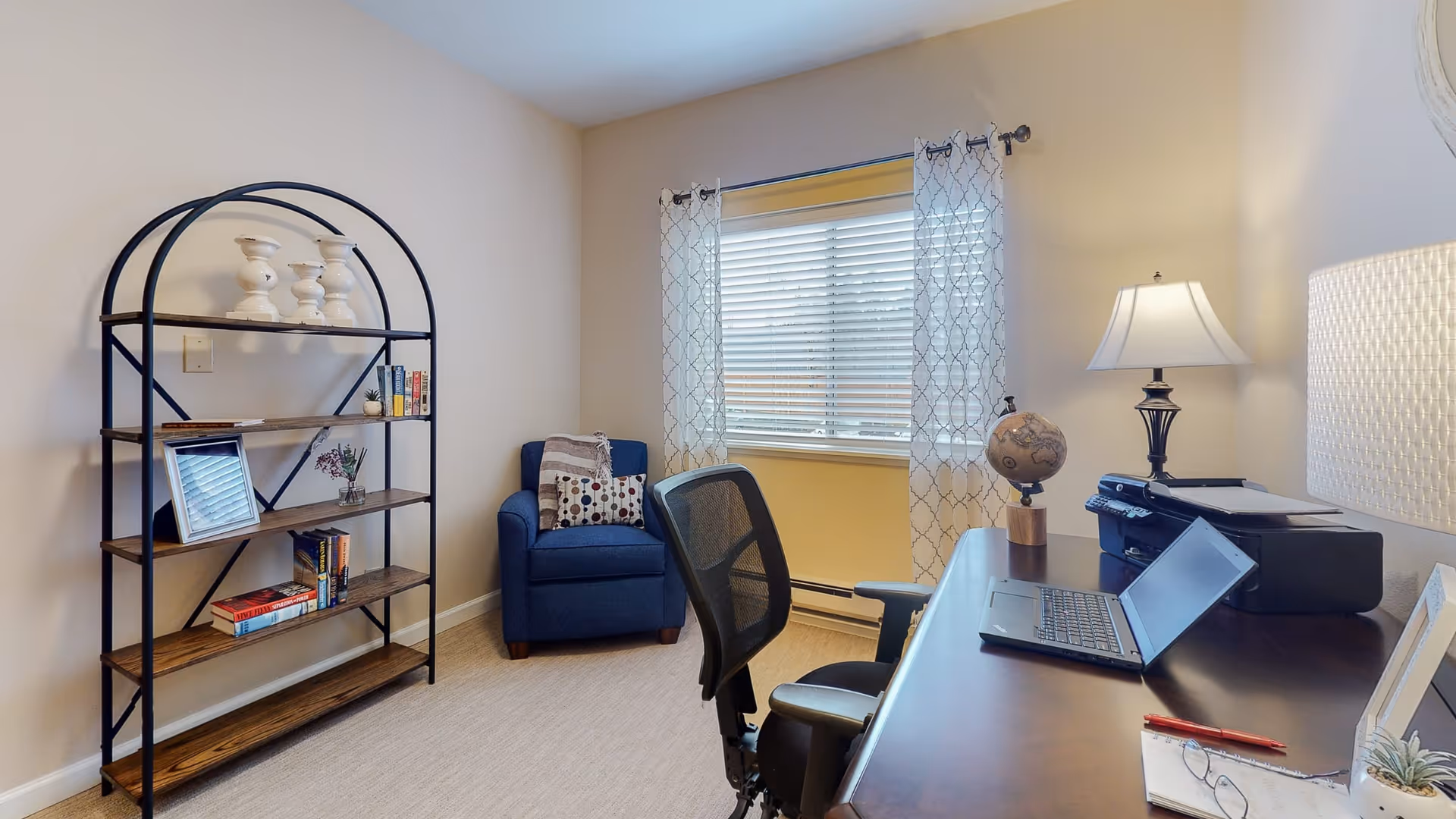 A cozy office or study room with a wooden desk holding a laptop, printer, globe, lamp, and some stationery. There is a black mesh office chair in front of the desk. To the left, a blue armchair with patterned pillows is positioned near a window with sheer patterned curtains. A black metal and wood bookshelf with decorative items and books stands against the wall.