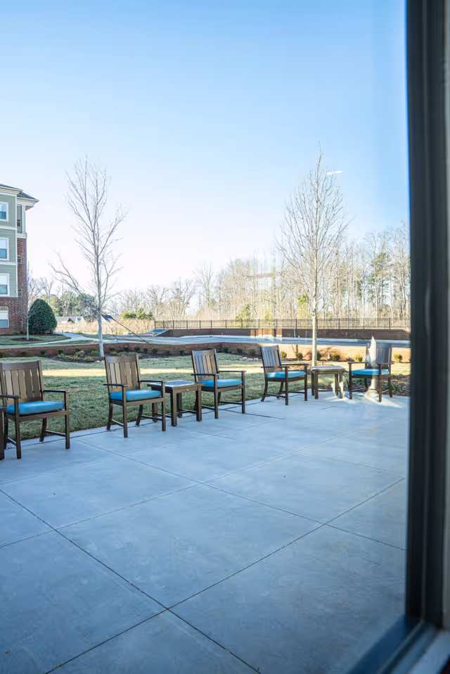 Outdoor patio area with six wooden chairs with blue cushions arranged around two small tables on a concrete surface, with grass, trees, and a building visible in the background under a clear blue sky.