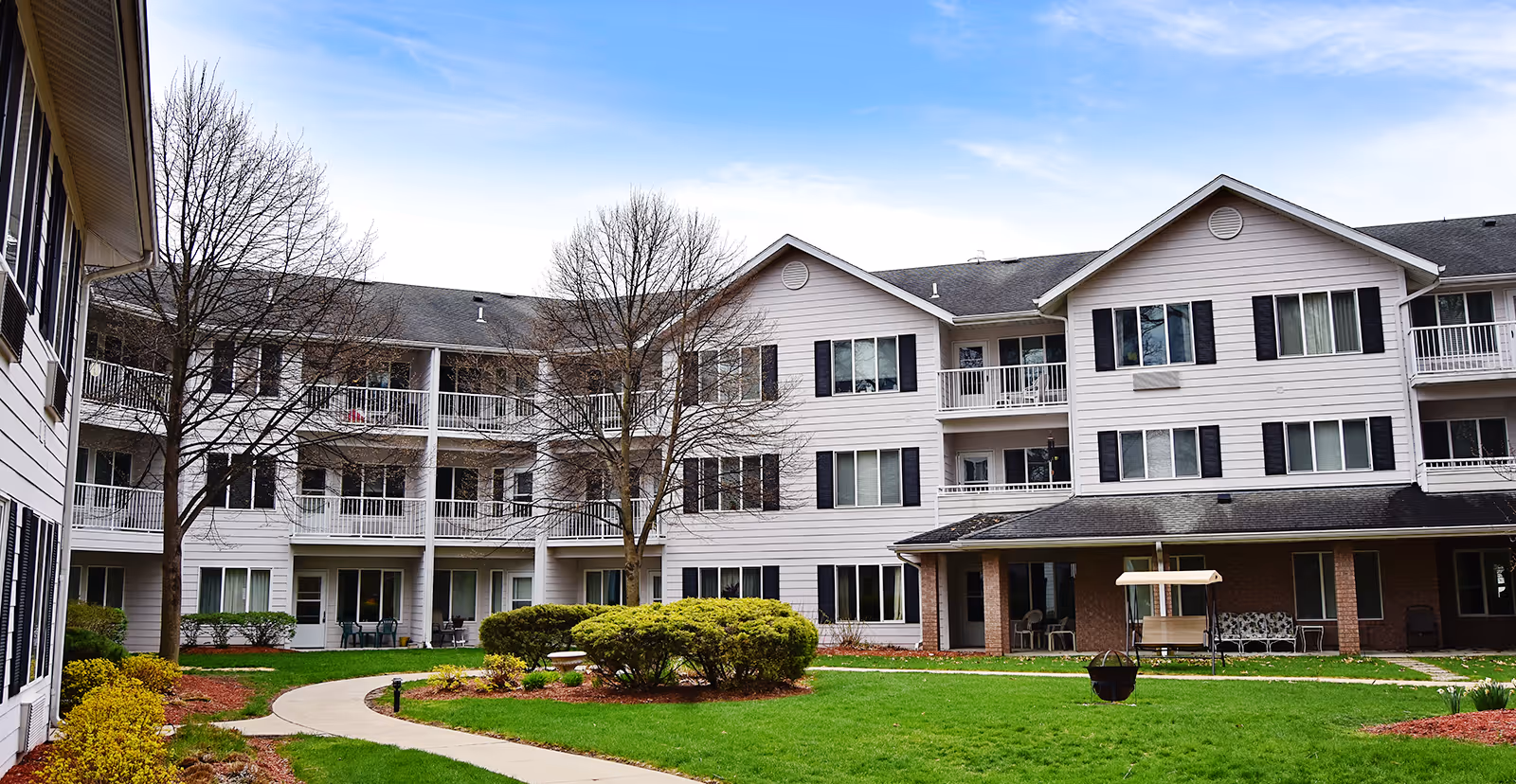 Exterior view of a three-story senior living facility building surrounding a landscaped courtyard with green grass, bushes, and leafless trees under a partly cloudy sky.