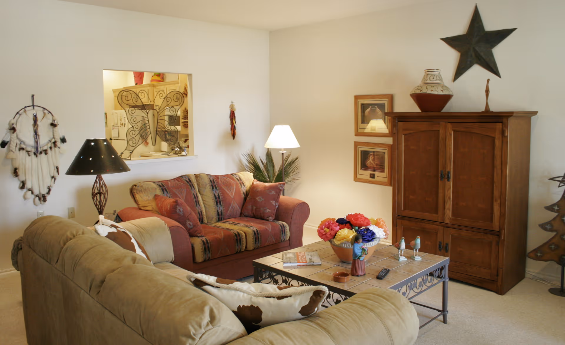 A cozy living room with two sofas, one beige and one patterned in red and brown tones. A coffee table with decorative items and a remote control sits between the sofas. There is a wooden cabinet against the wall with a large star decoration above it, two framed pictures, and a lamp beside the patterned sofa. The wall has a small window opening into another room, and there are various decorative items including a dreamcatcher and a metal tree sculpture.