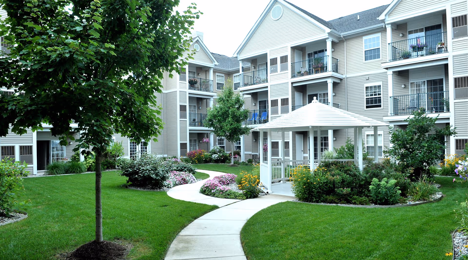 A landscaped courtyard with a winding concrete pathway, green grass, flower beds, and a white gazebo in the center. Surrounding the courtyard are multi-story residential buildings with balconies and windows.