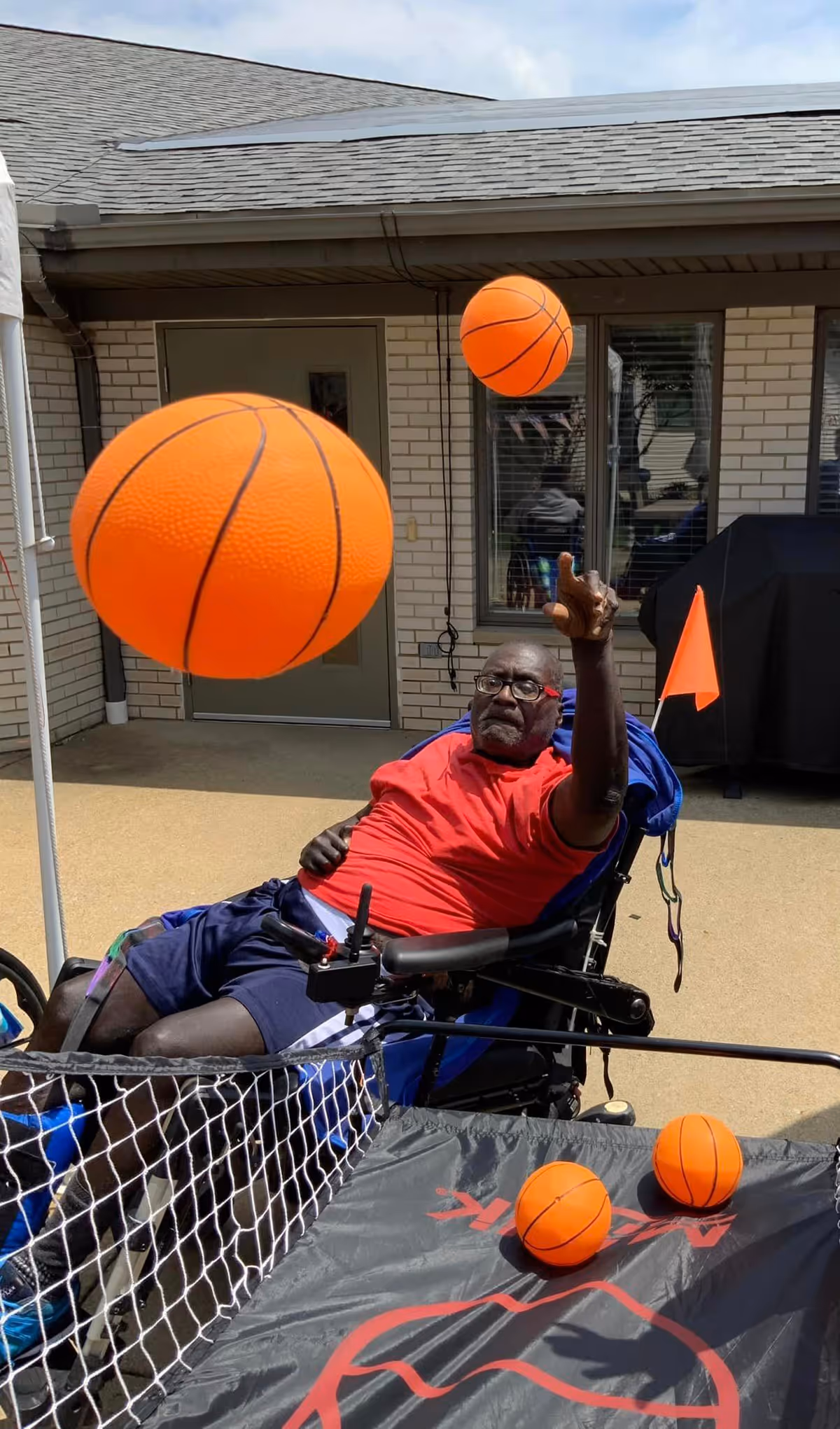 An elderly man in a wheelchair wearing a red shirt and blue shorts is playing a basketball tossing game outdoors at a senior care facility. There are three orange basketballs visible, one in mid-air and two on the game surface. The background shows a brick building with windows and a door.