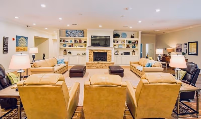 A spacious and well-lit living room with three tan leather armchairs in the foreground, two beige sofas facing each other, two dark ottomans, and a brick fireplace with a mounted flat-screen TV above it. Built-in shelves flank the fireplace, decorated with various items. Several table lamps provide warm lighting, and framed artwork adorns the walls.