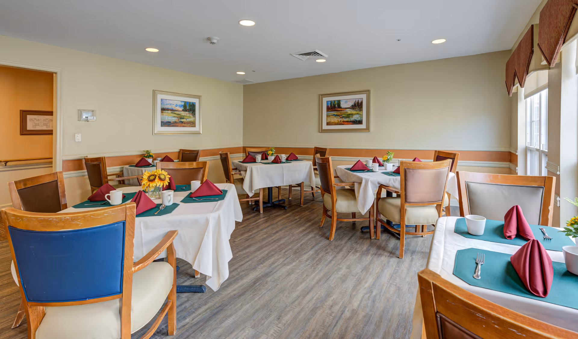 A dining room with several tables covered in white tablecloths and green placemats, each set with maroon folded napkins, white mugs, and silverware. The room has wooden chairs with cushioned seats, light-colored walls adorned with framed landscape paintings, and large windows with valances letting in natural light. The floor is a wood-like laminate.