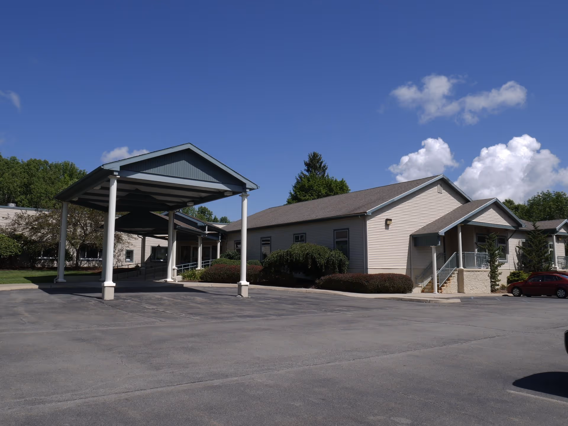Front exterior of a single-story senior care building with a covered drop-off canopy and parking lot under a blue sky.