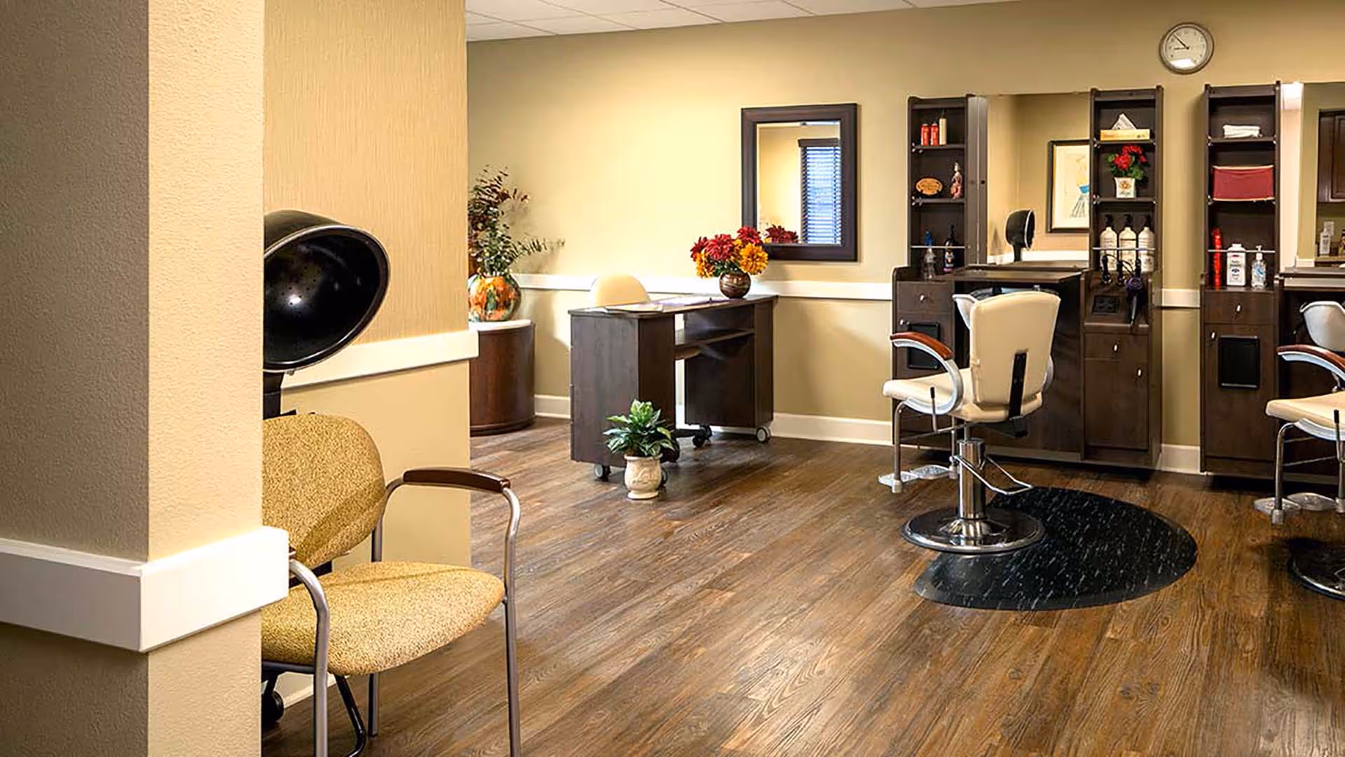 Interior of a hair salon area in a senior living facility with wooden flooring, beige walls, salon chairs, mirrors, and shelves stocked with hair care products. There is a small desk with a chair and decorative plants and flowers.