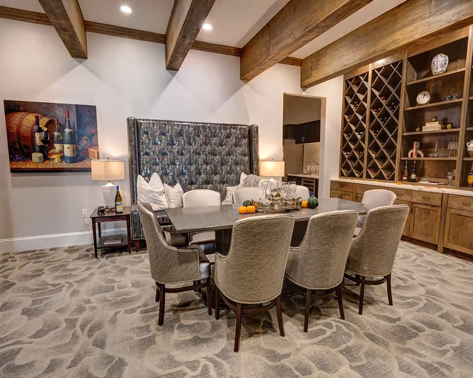 A dining room with a rectangular table surrounded by six upholstered chairs. The room features a tufted high-back bench against the wall, two table lamps on side tables, a painting of wine bottles and barrels, wooden ceiling beams, and built-in wooden shelves and wine racks filled with bottles and decorative items.