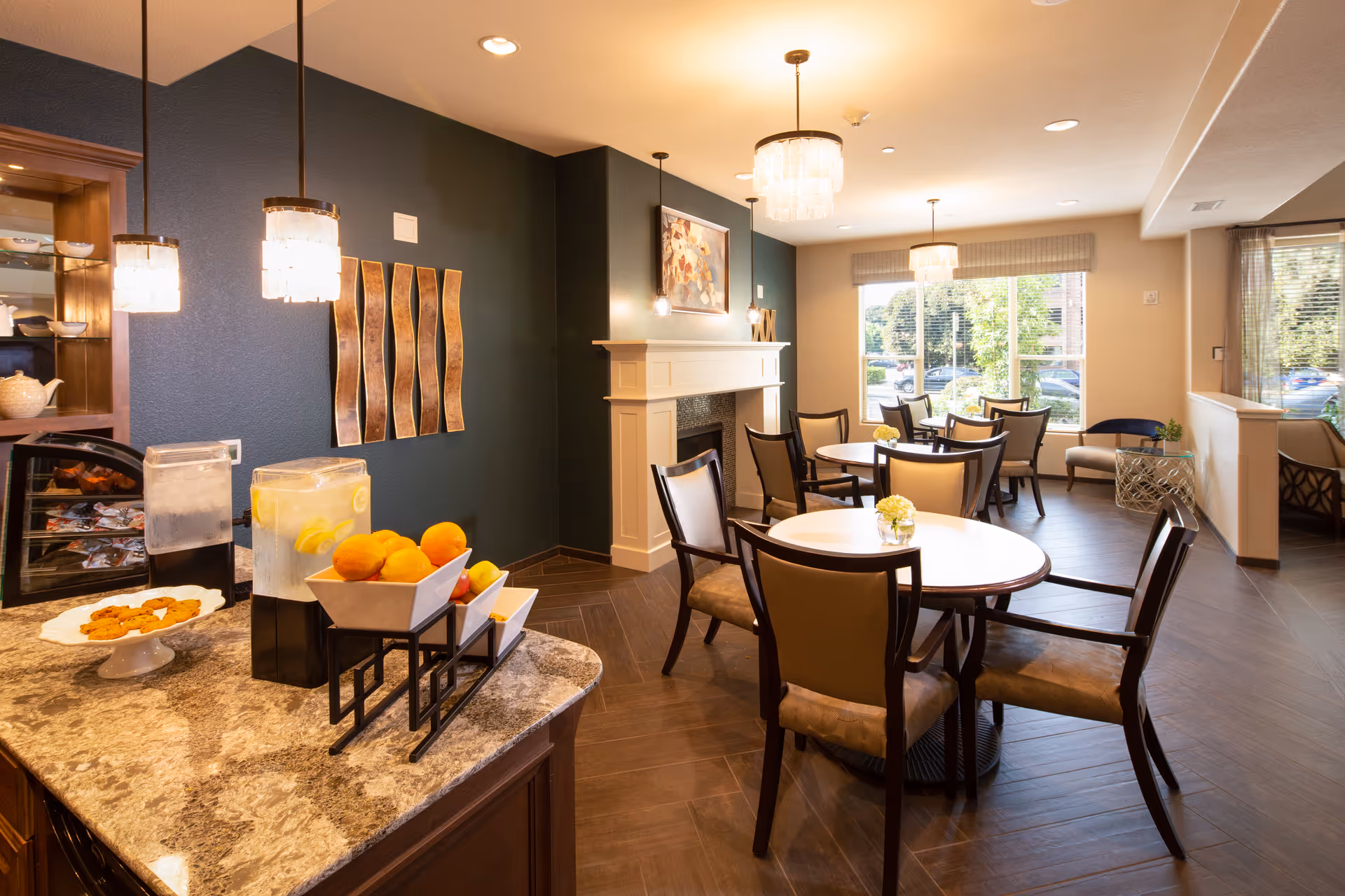 Well-lit dining room with round tables and chairs, a countertop beverage station with lemons, and large windows overlooking greenery.
