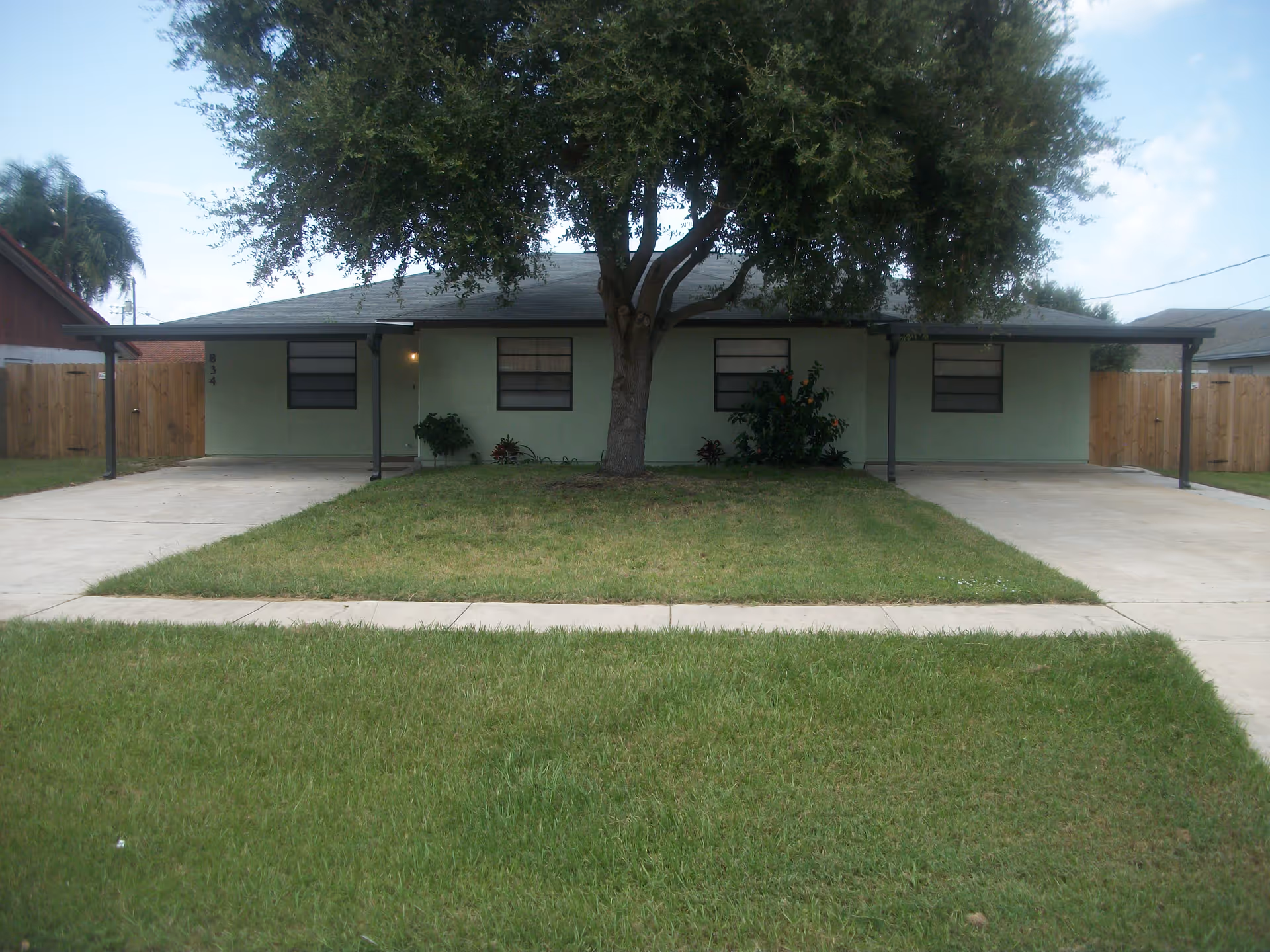 Single-story green building with a large front tree, twin carports and a grassy front lawn.