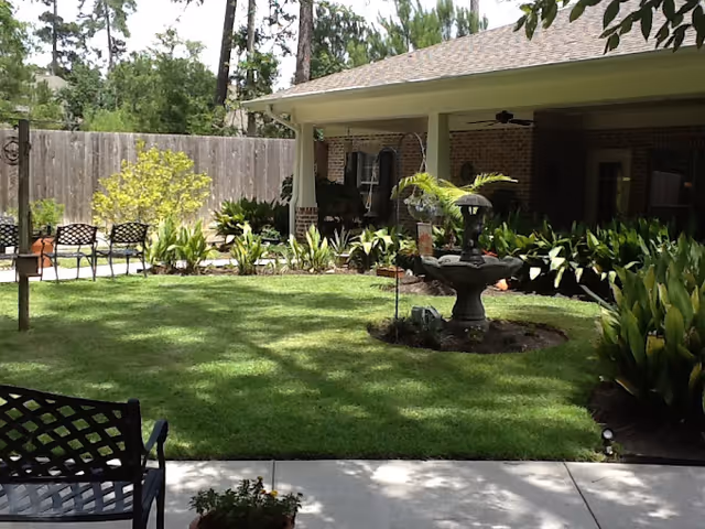 A well-maintained outdoor garden area with green grass, various plants, and a three-tiered water fountain in the center. There are several black metal chairs arranged along the edge of the garden near a wooden fence. A covered patio with brick walls and ceiling fans is visible in the background.