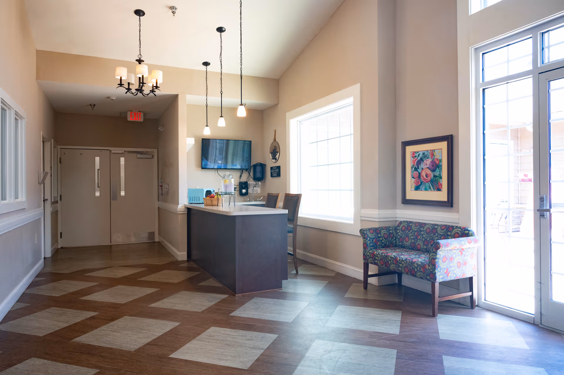 Interior view of a senior living facility reception area with a dark wood reception desk, two chairs, a wall-mounted TV, pendant lights, a floral patterned loveseat, a framed floral painting, large windows, and double glass doors leading outside.