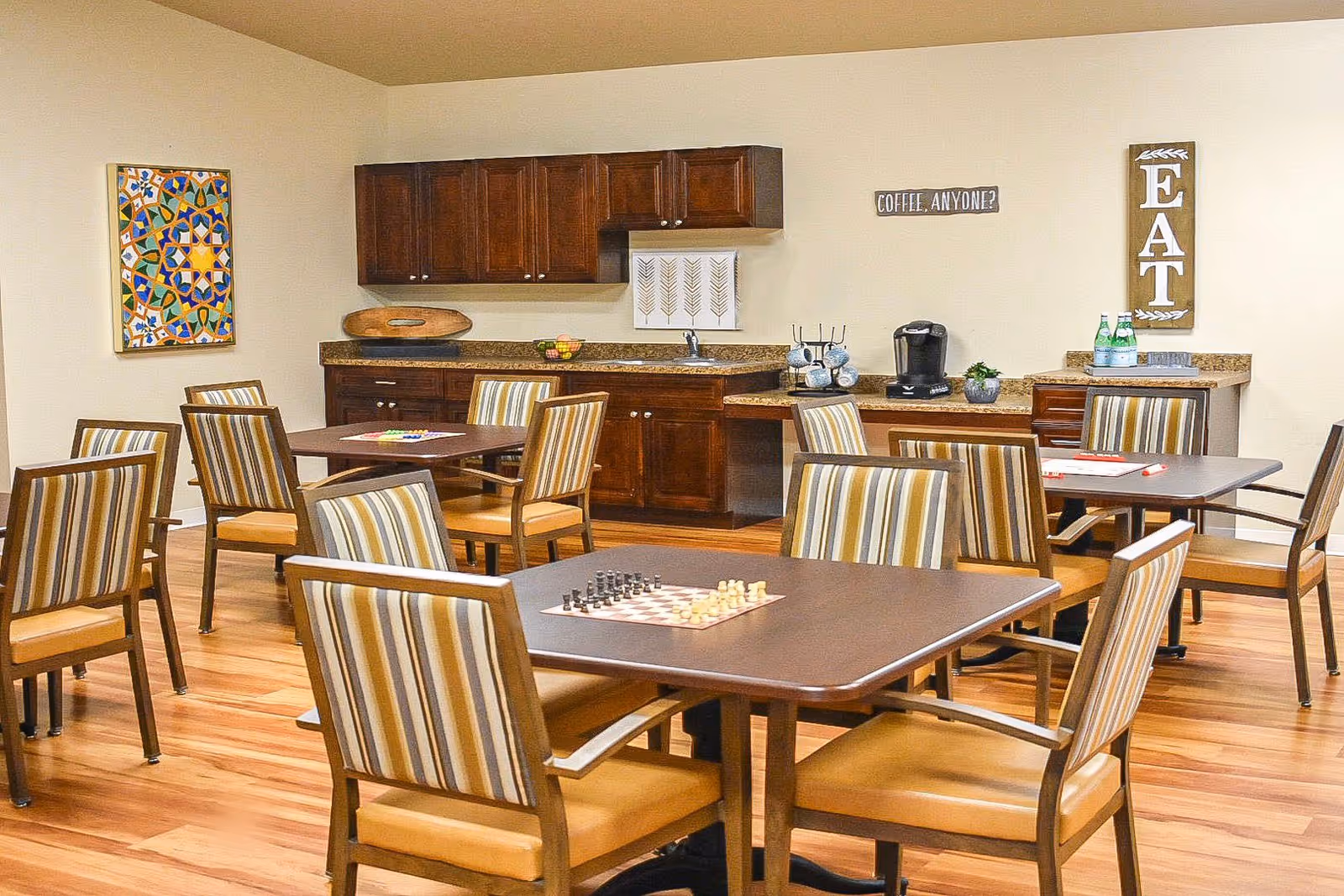 A communal dining and activity room with several tables and chairs featuring striped upholstery. One table has a chessboard set up. The room has wooden flooring, a kitchenette with dark wood cabinets, a coffee maker, and decorative wall signs including one that says 'COFFEE, ANYONE?' and another that says 'EAT'.